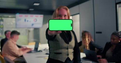 Horizontal smartphone with green screen held toward camera by businesswoman in office,