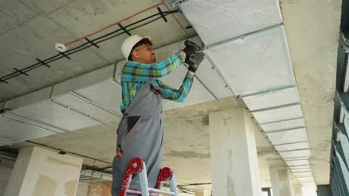 Construction Worker Drilling Ceiling Panel on Ladder