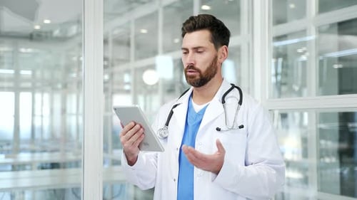Confident doctor in white coat talking on video call using digital tablet in hospital office.