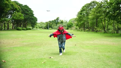 Happy girl are playing superhero. Kid run across green field in red cloak