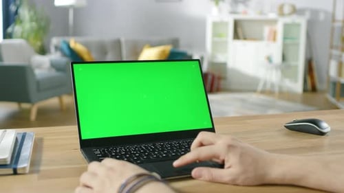 Man Uses Laptop with Green Mock-up Screen While Sitting at the Desk in His Cozy Living Room.