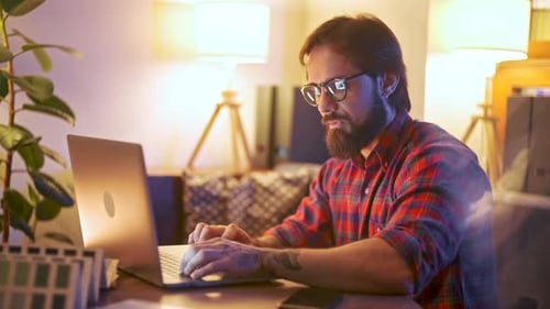 Focused Bearded Professional Working on Laptop in Cozy Office Hand Holding Smartphone with Green