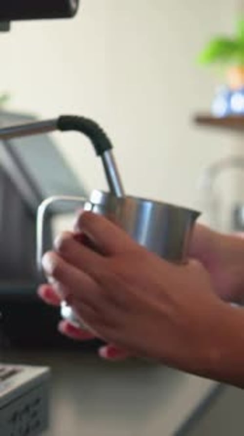 Vertical video: Starting steam wand, hands frothing milk in pitcher at cafe for latte, denim sleeve