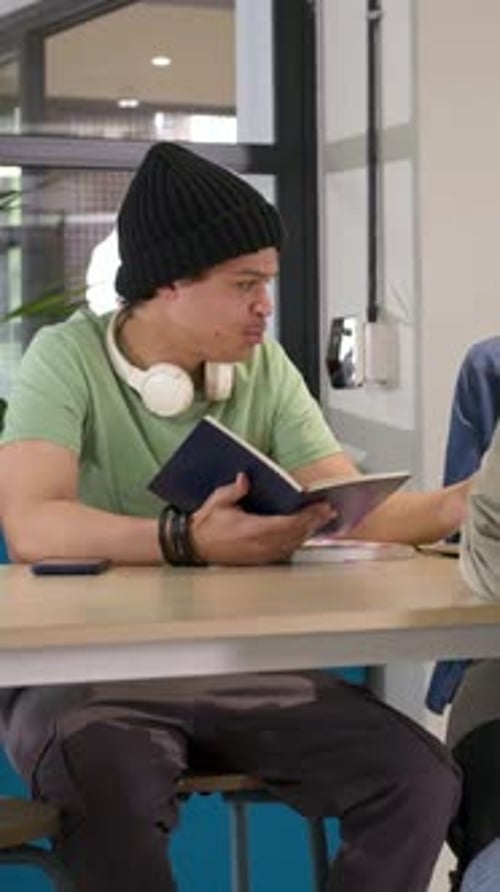 Vertical video: Smiling student discussing notes in lounge, pointing at navy notebook with pencil