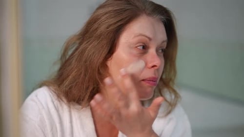 Woman Applying Moisturizer to Face in Mirror