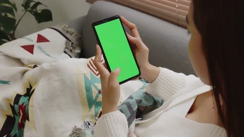 Young Asian woman using smartphone on couch with blank green screen chroma key.