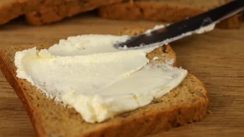 Frischkäse mit einem Messer auf einem Stück Mehrkornbrot auf einem Holzschneidebrett verteilen.