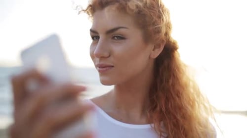 Happy young redhead woman taking selfie with smartphone on beach in summer