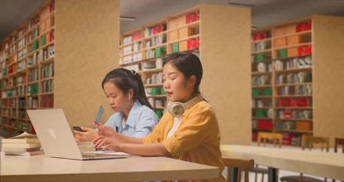 Student Use A Laptop And Writing Into The Notebook While Sitting With Her Classmate In The Library