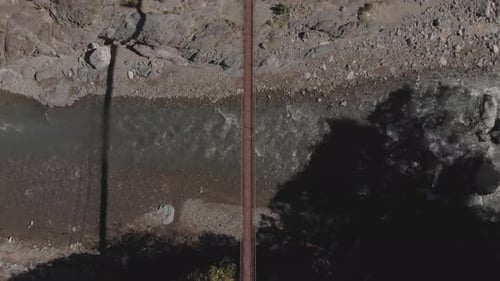 two people walking across a metal wire rope suspension bridge over rocky river water flowing down mo