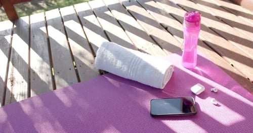 Towel, bottle with water, smartphone and headphones lying on yoga mat on sunny terrace, slow motion