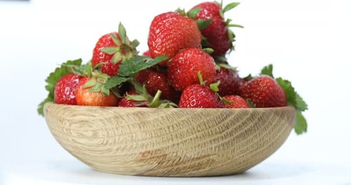 Many fresh ripe strawberries with green leaves in bowl on white background, rotating