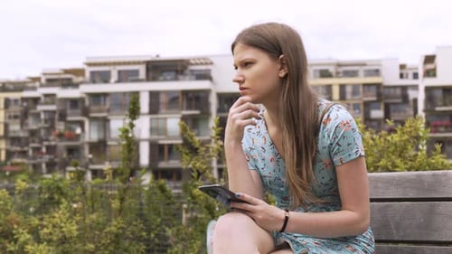 A Beautiful Young Caucasian Woman Works on a Smartphone As She Sits on a Bench in an Urban Area