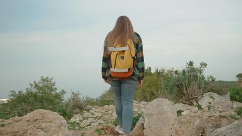 A young, long-haired woman with a backpack navigates through rocks on a high mountain, overlooking.