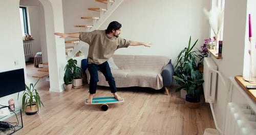 A Brunette Guy in a Green Tshirt is Trying to Stand on an Unstable Plank and Cylinder Structure to