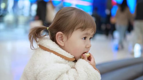 Side View Of A 3-Year-Old Toddler Girl Sitting Eating Bread Inside A Shopping Mall. - Selective Focu