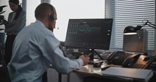 Male Financial Trader in Headset Works on Computer Monitors Stocks Takes Notes