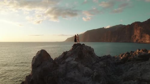 Young couple stands on a large volcanic rocky formation by the sea, holding hands and watching the d