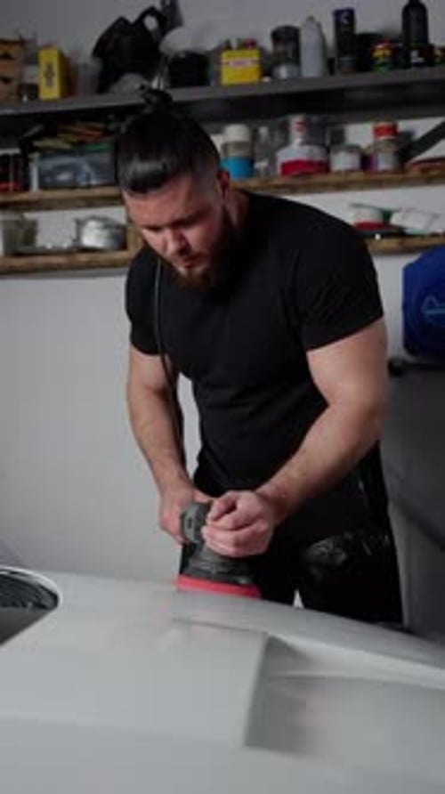 A Man in a Black Tshirt and a Pigtail on His Head Polishes a White Car in a Car Studio