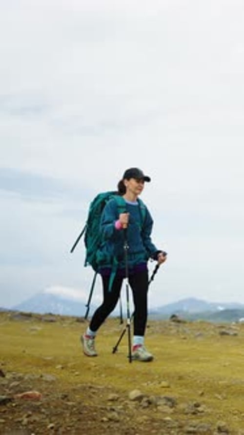 Solo Female Hiker Trekking Across Barren Mountain Trail with Backpack and Poles Under Overcast Sky