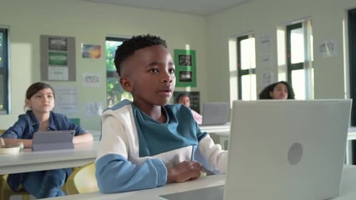 African American boy at desk raising hand, responding to teacher, using silver laptop