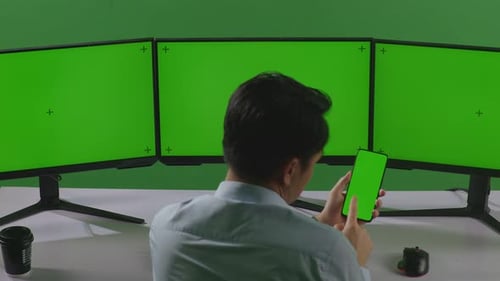 Asian Man Using Mobile Phone And Multiple Computer Monitor With Mock Up Green Screen In Green Screen