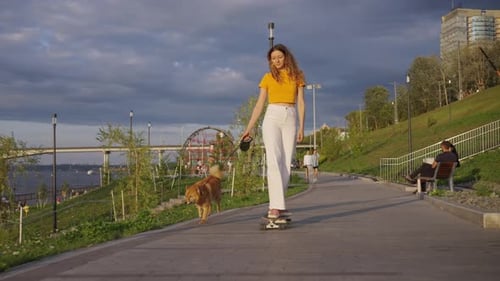 Young Woman Skateboards with Her Dog Along a Riverside Promenade at Dusk