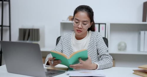 Asian adult woman sitting indoor reading green book while holding pen and thinking at modern home