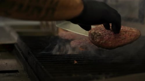 Chef uses spatula to turn burger patty on grill, three hamburgers cook sizzle over fire, 4K