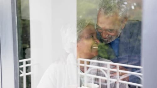 Happy senior biracial couple looking through window and embracing, unaltered, in slow motion