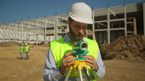 Joven caucásico barbudo con chaleco amarillo y casco protector utilizando la estación total en una construcción