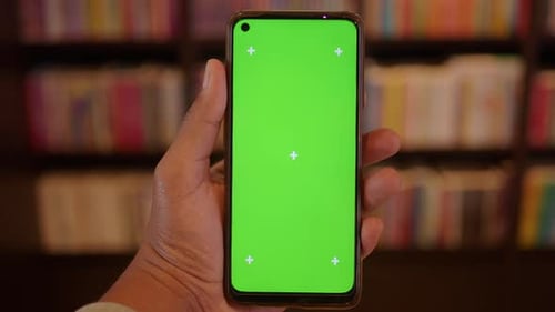 Hand Holding Smartphone with Green Screen in Cozy Library
