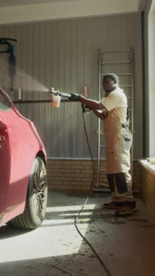 Vertical of Worker Washing Red Car with Hose in Garage