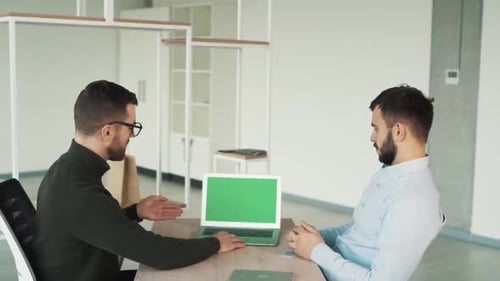 Two young men are sitting at the table and looking at the green screen of the laptop