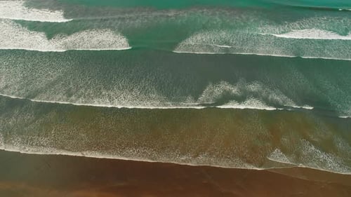 Aerial flight over ocean waves forming rhythmic patterns along vast sandy coastline