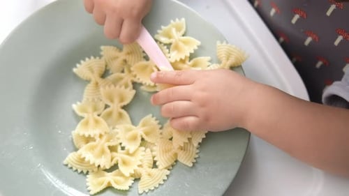 Top View Caucasian Baby Girl About 2 Years Old in Bib Eating Pasta From Plate Sitting High Chair