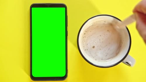 Smart Phone with Empty Green Screen Next to Coffee Cup Person Hand Stirring Sugar with Spoon Against