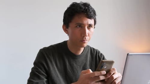 Eurasian man thinking about a text he's writing on his phone while working from home on his computer