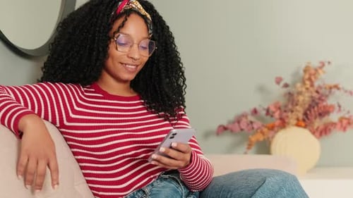 Happy woman sitting on sofa in living room texting sms using mobile phone