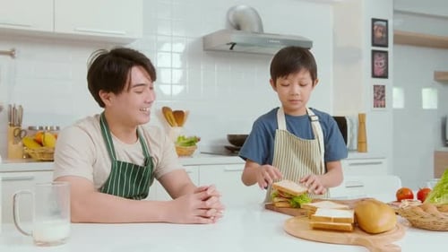Smiling Man Watches Boy Prepare Sandwich in Kitchen