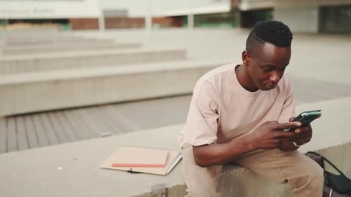 Young student sits outside of university, uses cellphone, writes on social networks