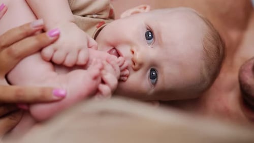 Grey-eyed infant holding and biting his tiny feet. Father holds his little son in hands. Close up.