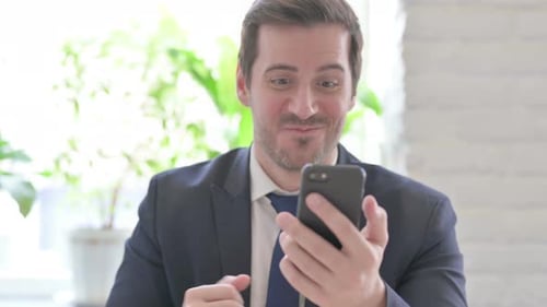 Young Businessman Celebrating Online Success on Smartphone in Office