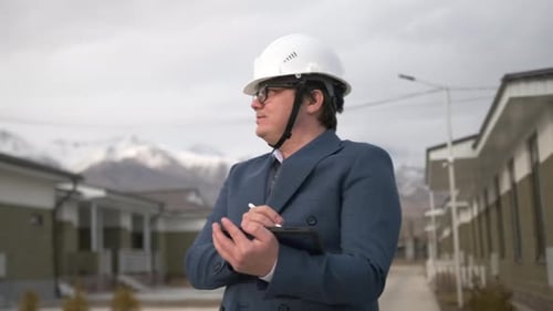 An architect takes notes on a tablet, wearing a suit and helmet against the background of houses