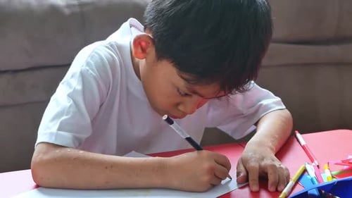 Junger asiatischer Junge konzentriert sich aufs Zeichnen und Schreiben und hat farbenfrohe Malutensilien parat
