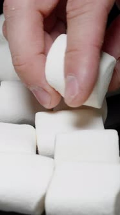 Vertical video. Male Hand Examining a White Marshmallow, Many Marshmallows Scattered on the Table