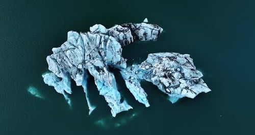 Top-down drone view of sculpted blue icebergs floating in a dark glacier lagoon, with finger-like