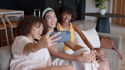 Amigas afro-americanas no sofá, mulher deixada estendendo o smartphone verificando selfies, reagindo