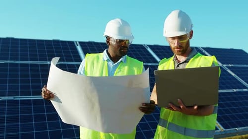 Serious Men Use Laptop and Project Plan on the Solar Farm in Special Uniform Standing Discuss the