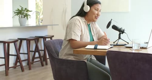 Femme mettant des écouteurs, parlant dans un microphone, écrivant des notes sur son espace de travail à domicile pour une réunion